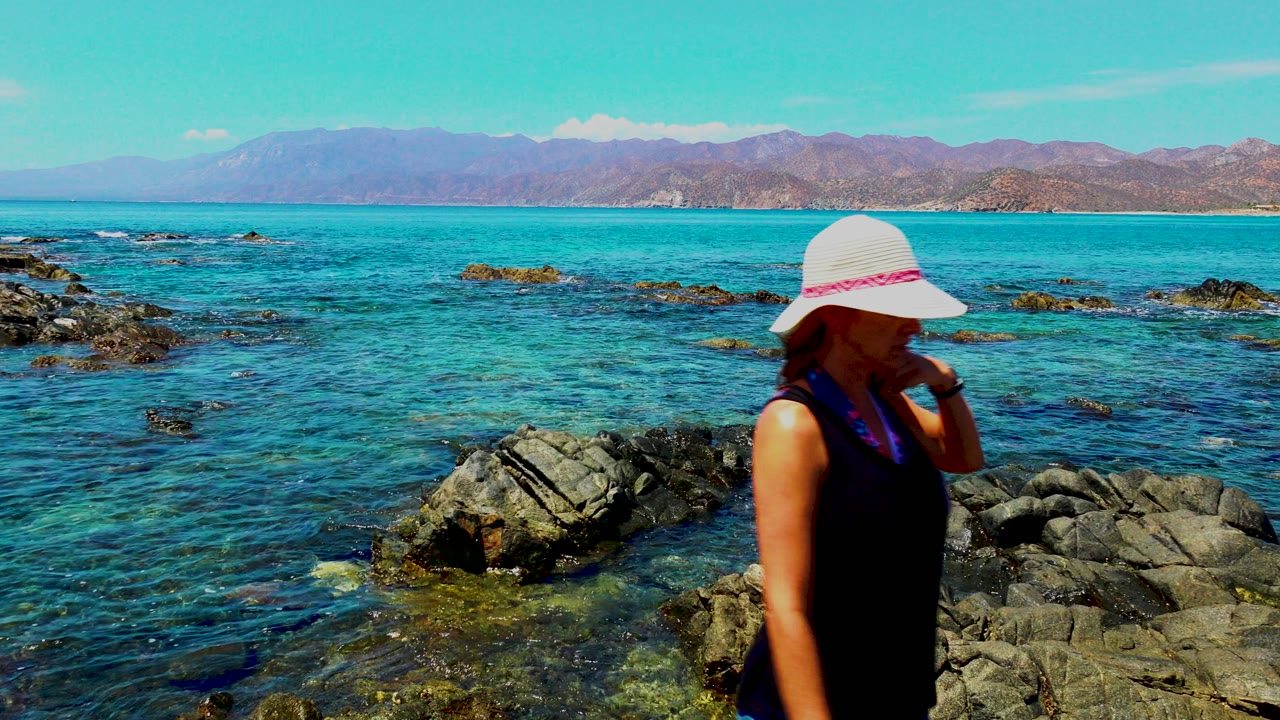 At El Sargento La Ventana Mexico, the woman has moved toward the right edge while rocks, shallow blue-green water, and a mountain horizon fill the frame.