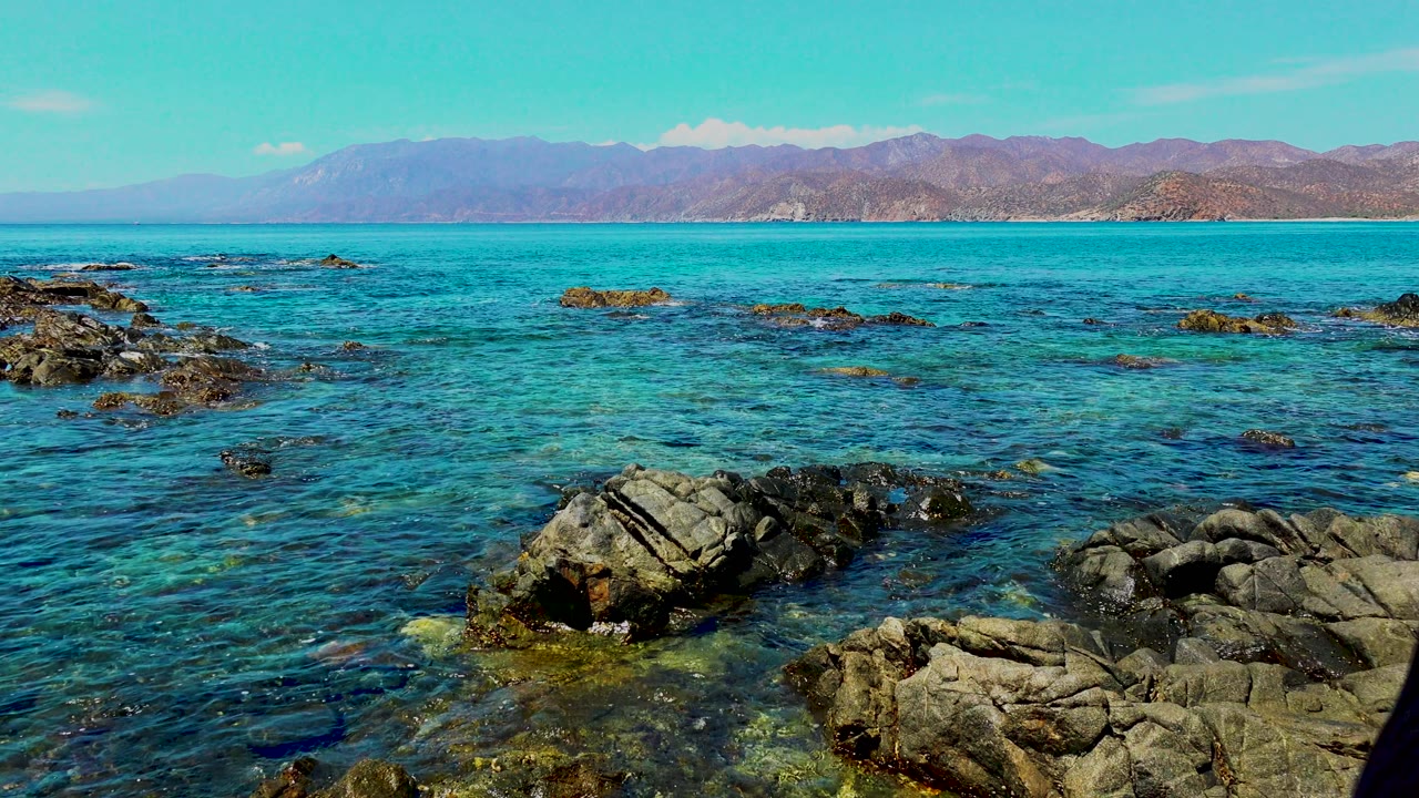 At El Sargento La Ventana Mexico, only a narrow edge of the woman remains at the far right as turquoise water, scattered rocks, and distant mountains dominate the view.