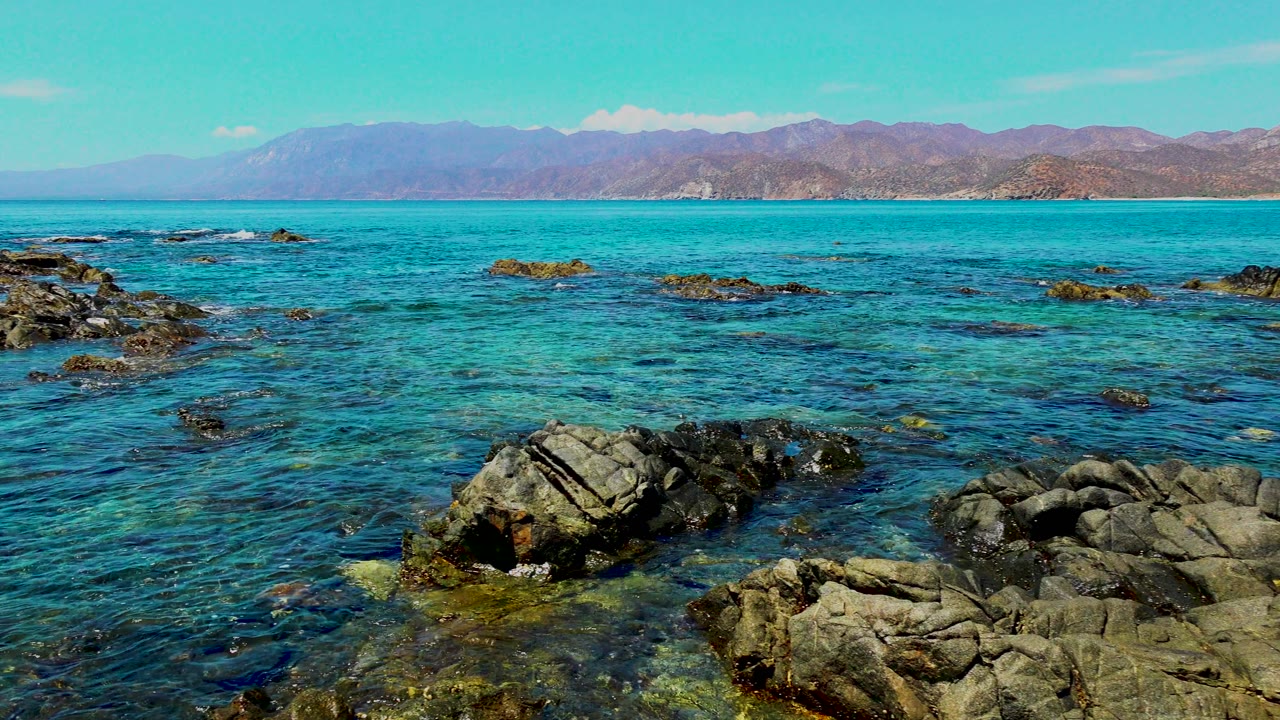 Rocky shoreline with clear turquoise ocean water and mountains in the distance.