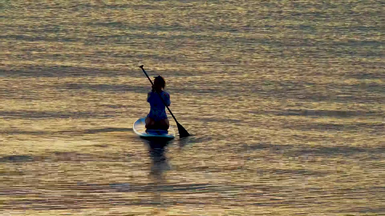 A person on a blue stand-up paddleboard in El Sargento La Ventana, Mexico, holds a paddle upright over golden rippled water at the start of the clip.