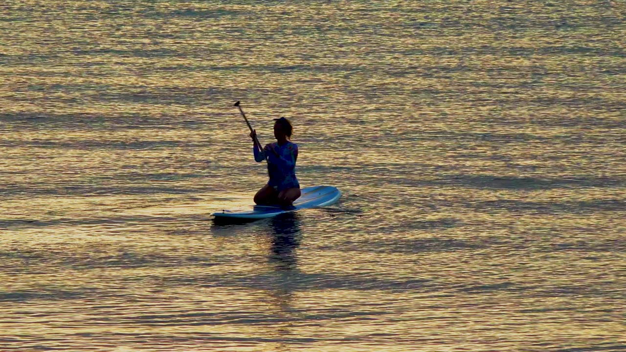 A distant person on a stand-up paddleboard in El Sargento La Ventana, Mexico, pauses with the paddle raised as warm reflections spread across the water.