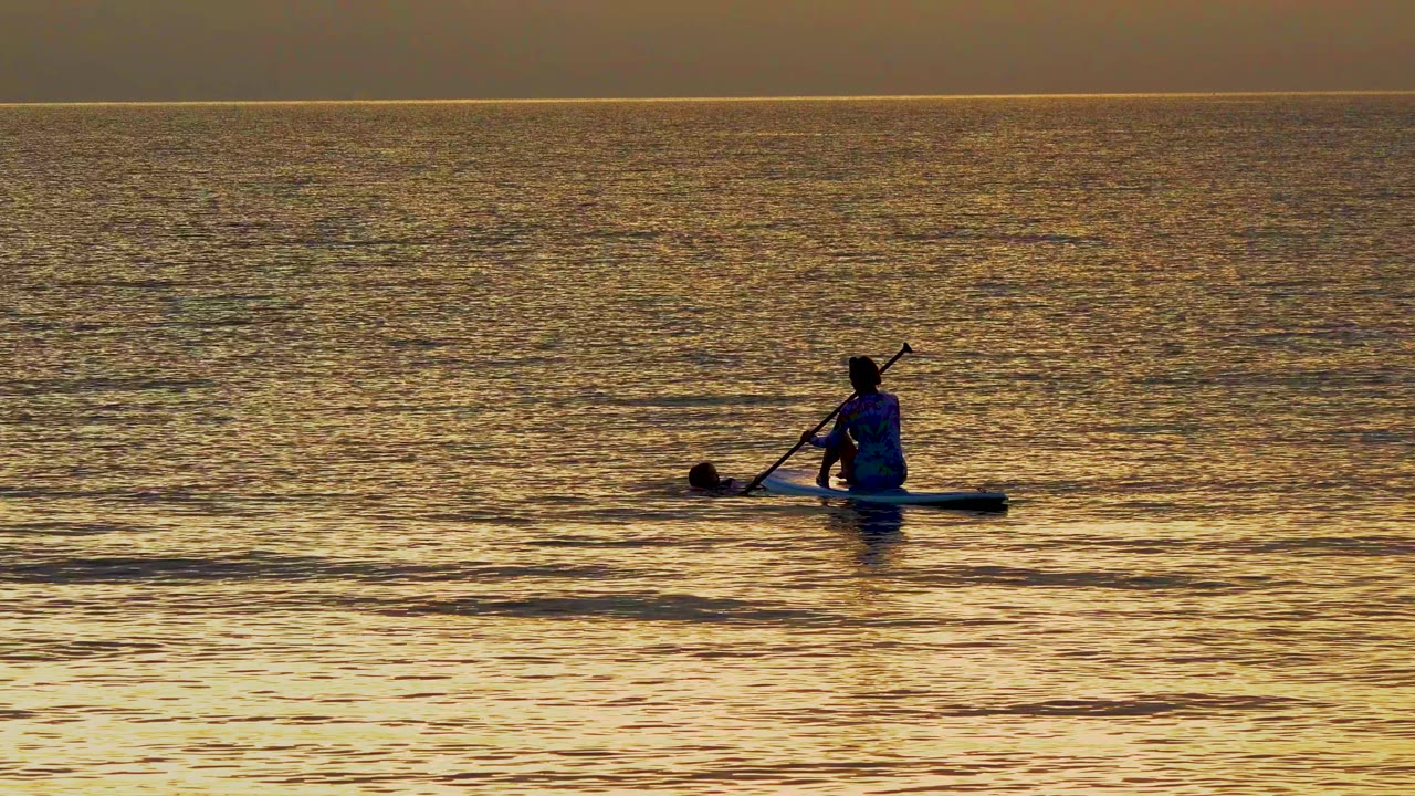 Paddleboarder with paddle angled down on calm sunset water.