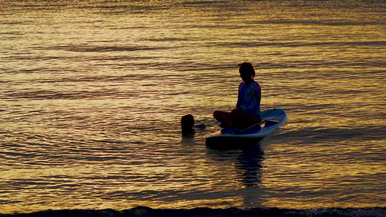 A silhouetted paddleboarder sits on a board near a swimmer in golden sunset water in El Sargento La Ventana, Mexico, with a small shore ripple at the bottom edge.