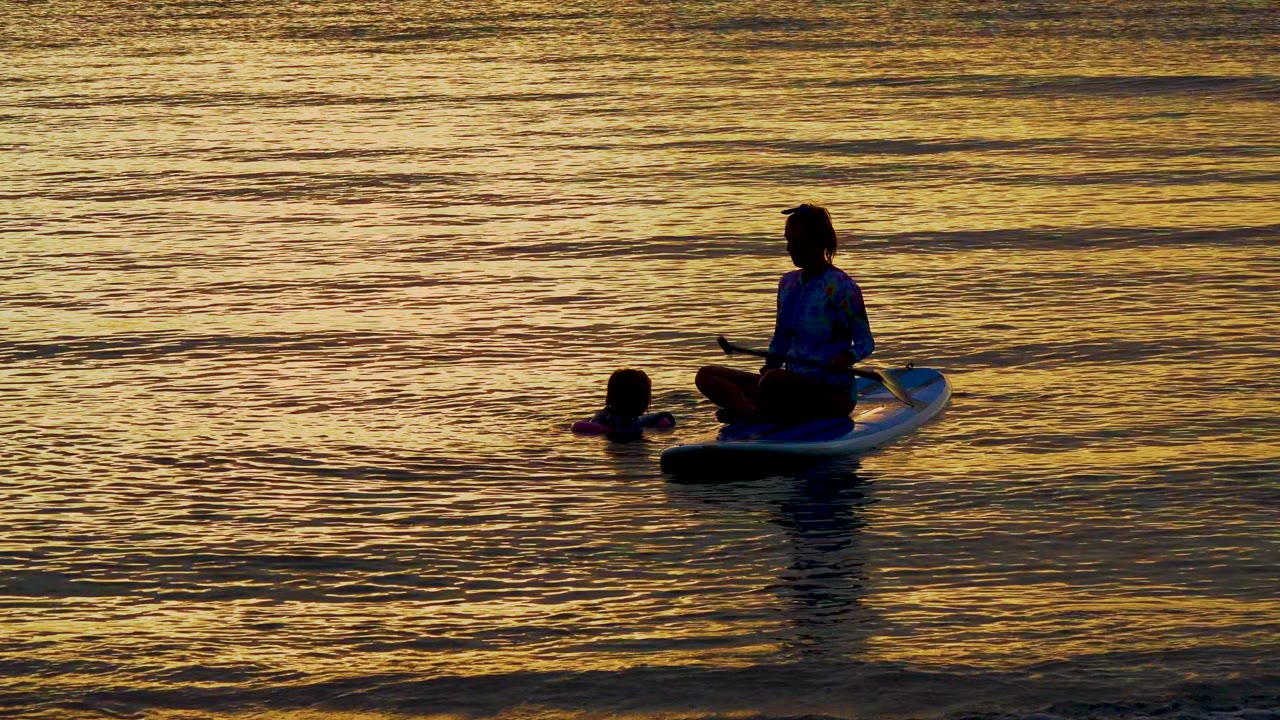 The paddleboarder remains seated on the board in El Sargento La Ventana, Mexico, holding the paddle low as the swimmer's head stays visible in the water beside the board.
