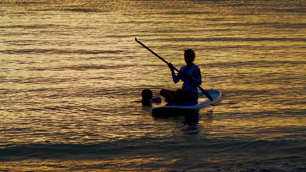 A silhouetted paddleboarder in El Sargento La Ventana, Mexico, raises the paddle diagonally while seated on the board, with the swimmer still close by in the gold-toned water.