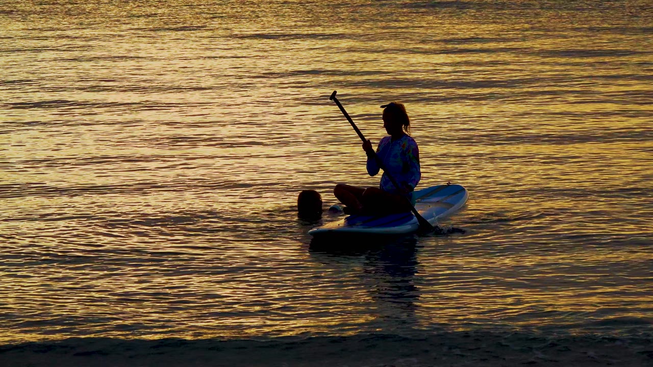 Paddleboarder sits with paddle upright near a swimmer in sunset water.