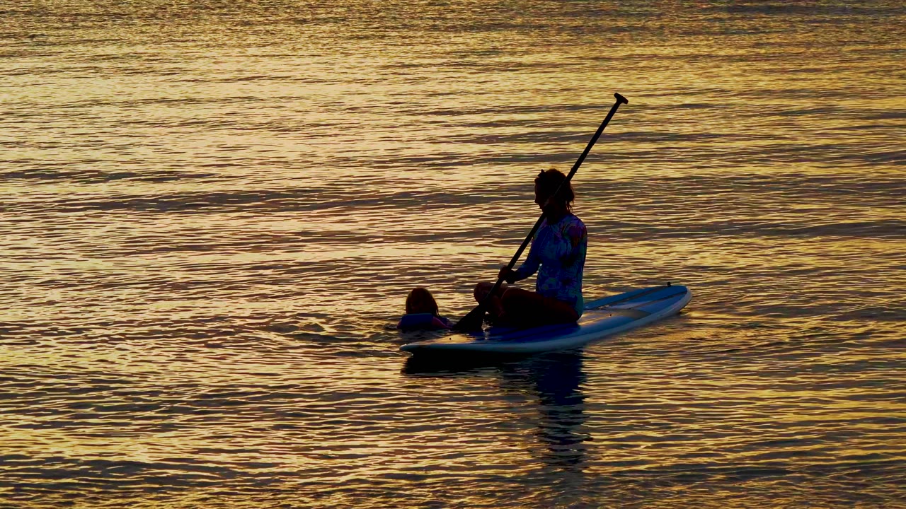 In El Sargento La Ventana Mexico, an adult sits on a paddleboard with a raised paddle while a child floats close to the board in sunset-colored water.