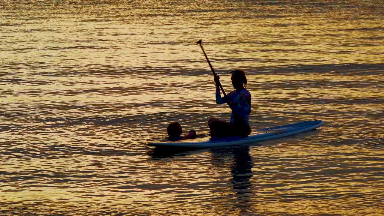 In El Sargento La Ventana Mexico, the adult raises the paddle nearly upright while the child floats ahead of the board on the textured sunset water.