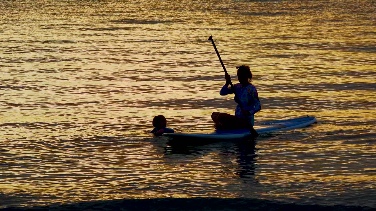 In El Sargento La Ventana, Mexico, a silhouetted person sits on a paddleboard with a raised paddle while a swimmer floats nearby in gold sunset water.