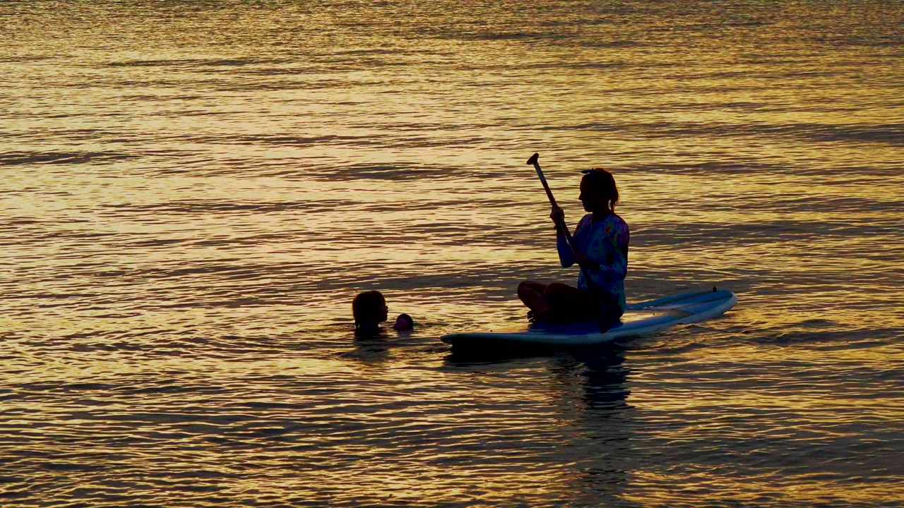Paddleboarder holds paddle upright near swimmer at sunset