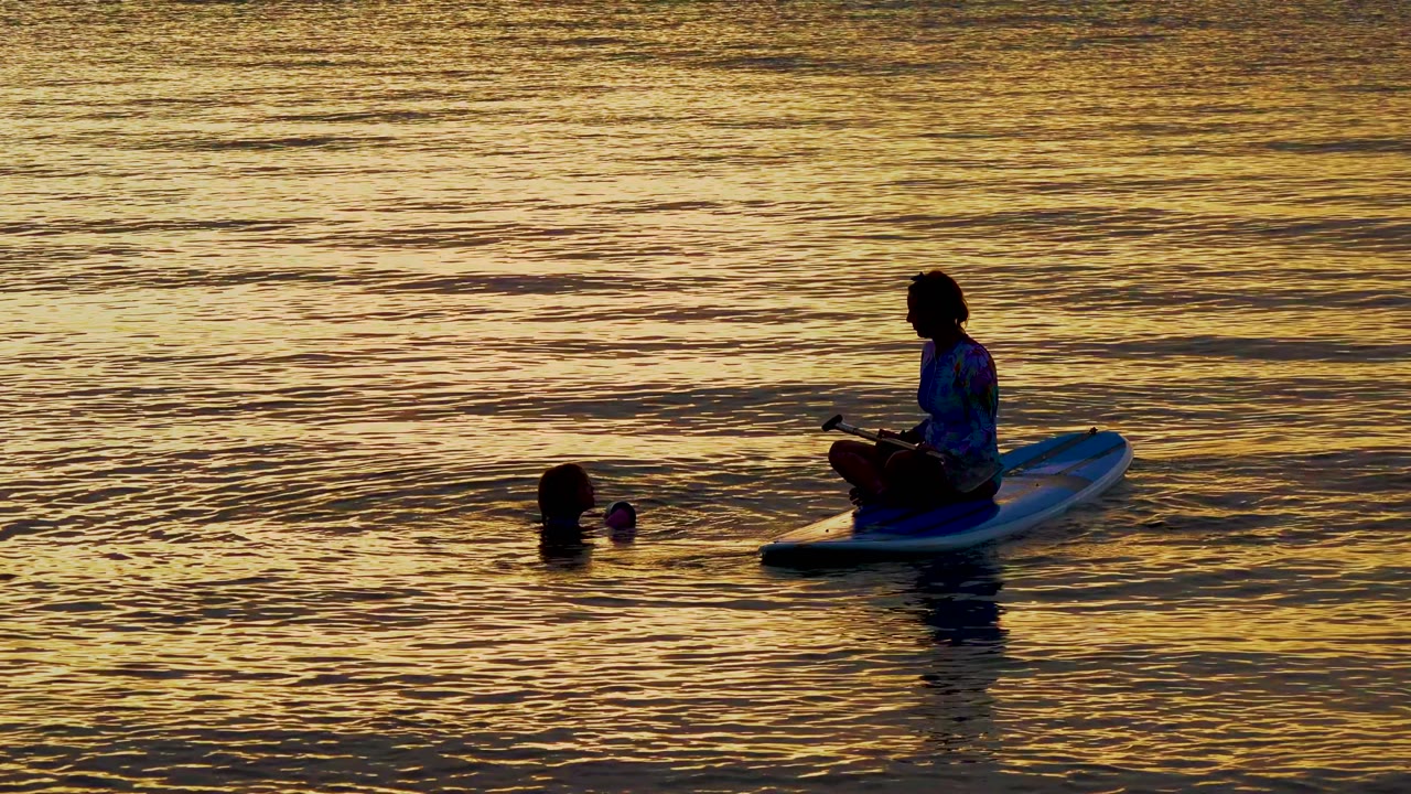 In El Sargento La Ventana, Mexico, the silhouetted paddleboarder sits farther right on the board while the swimmer and a small floating object sit left of the board in sunset reflections.