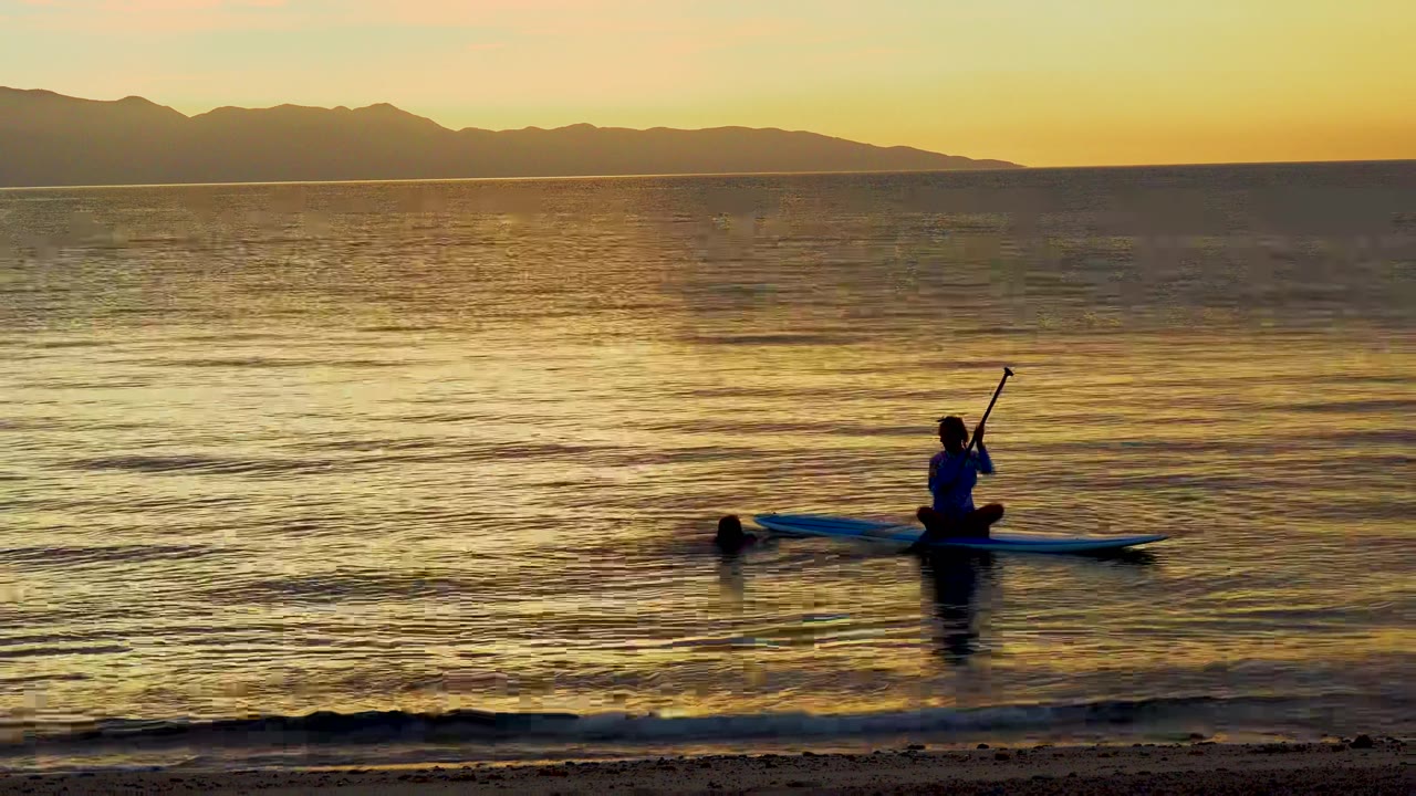 At 0:00 in El Sargento La Ventana, Mexico, a paddleboarder sits near a swimmer on golden ocean water with sandy beach foreground and mountains on the horizon.