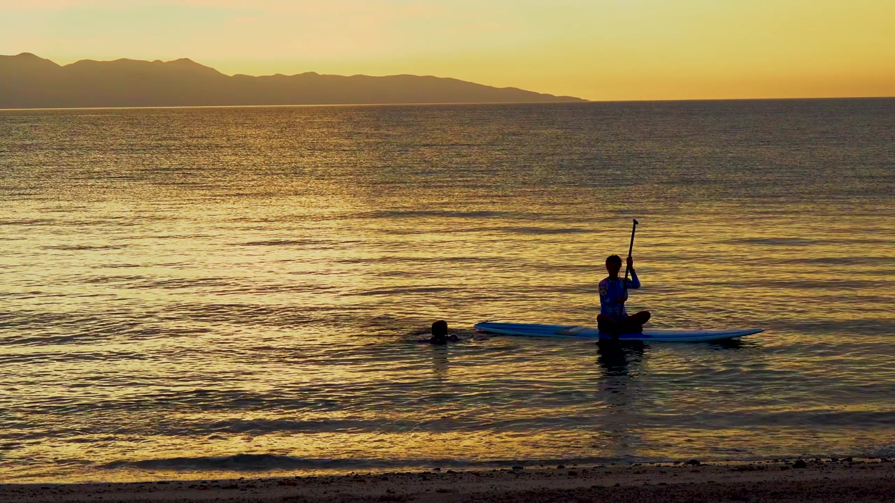 At 0:02 in El Sargento La Ventana, Mexico, the paddleboarder is positioned to the right of the swimmer, with the board parallel to the horizon on reflective sunset water.