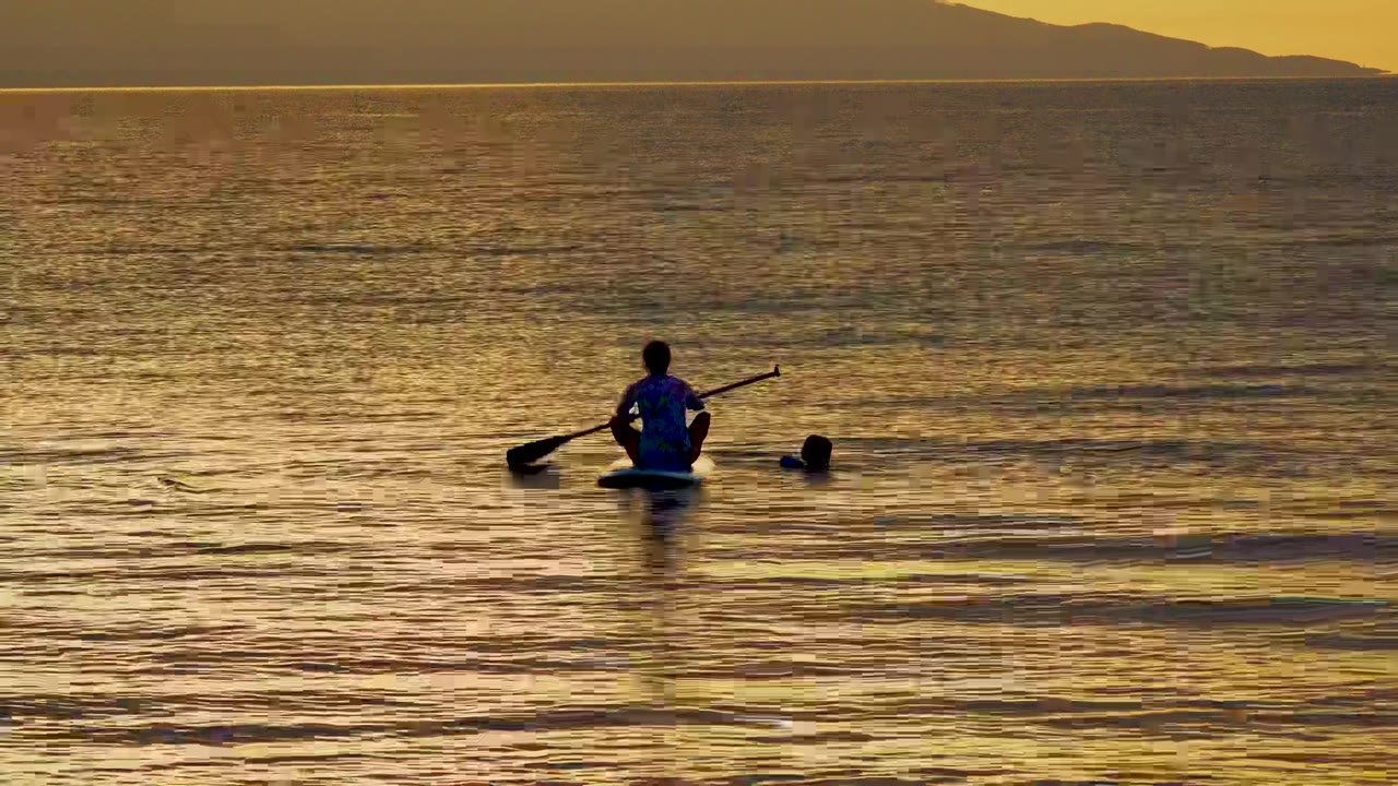 A person sits on a water float holding a paddle in golden sunset water at El Sargento La Ventana, Mexico, with a second head visible nearby and mountains on the horizon.