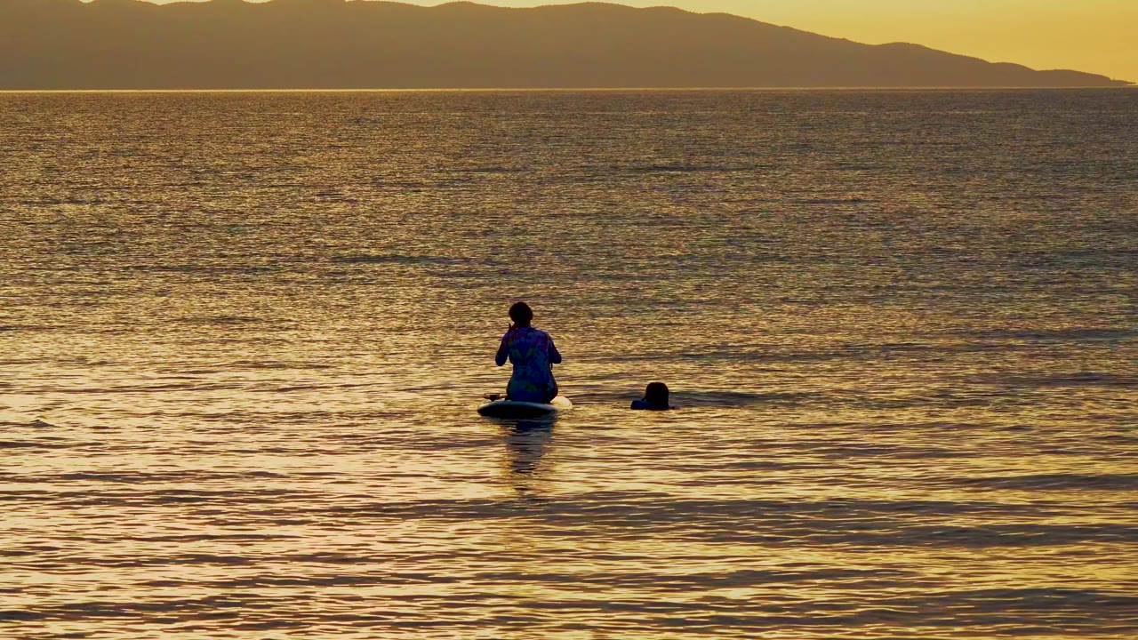 A person on the float raises their hands near their face in the sunset-lit water at El Sargento La Ventana, Mexico, with a second head visible to the right and mountains behind.