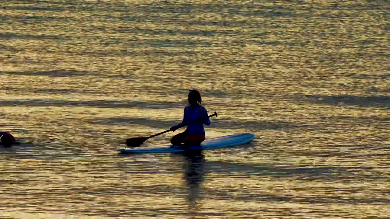In El Sargento La Ventana, Mexico, a silhouetted paddleboarder sits on a blue board while a swimmer is partly visible at the left edge in golden water.