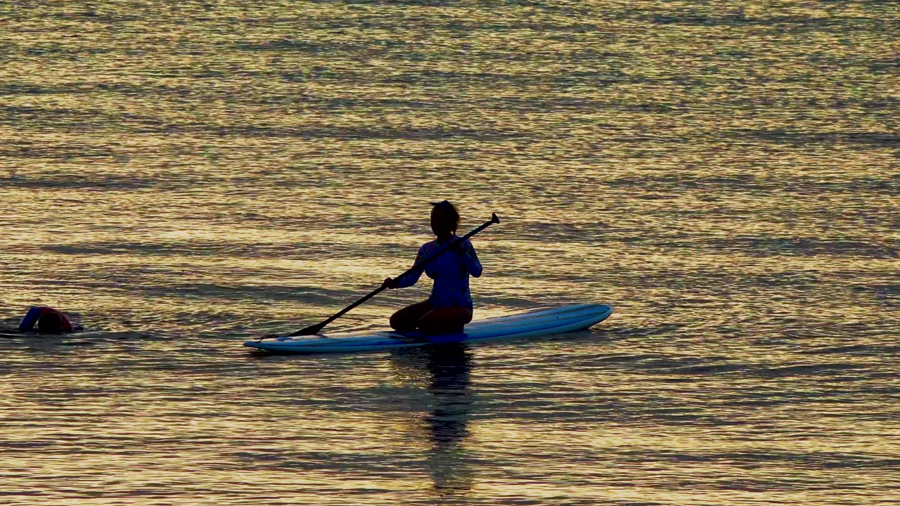 The paddleboarder sits upright with the paddle angled into the water while a swimmer surfaces in the surrounding golden reflections in El Sargento La Ventana, Mexico.