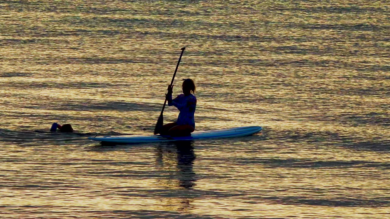 At 0:00 in El Sargento La Ventana Mexico, a silhouetted person sits on a blue paddleboard with a paddle raised while a swimmer is low in the water to the left.