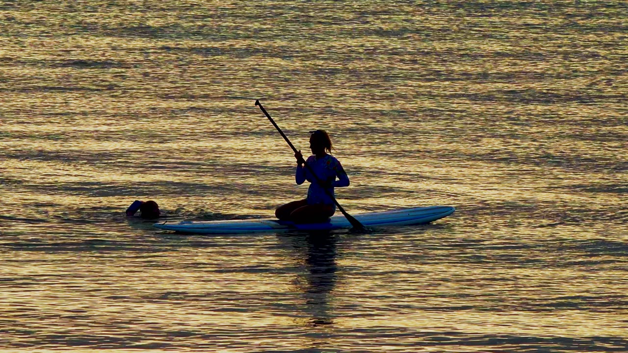 Paddleboarder holds a paddle while swimmer stays nearby in reflective water.