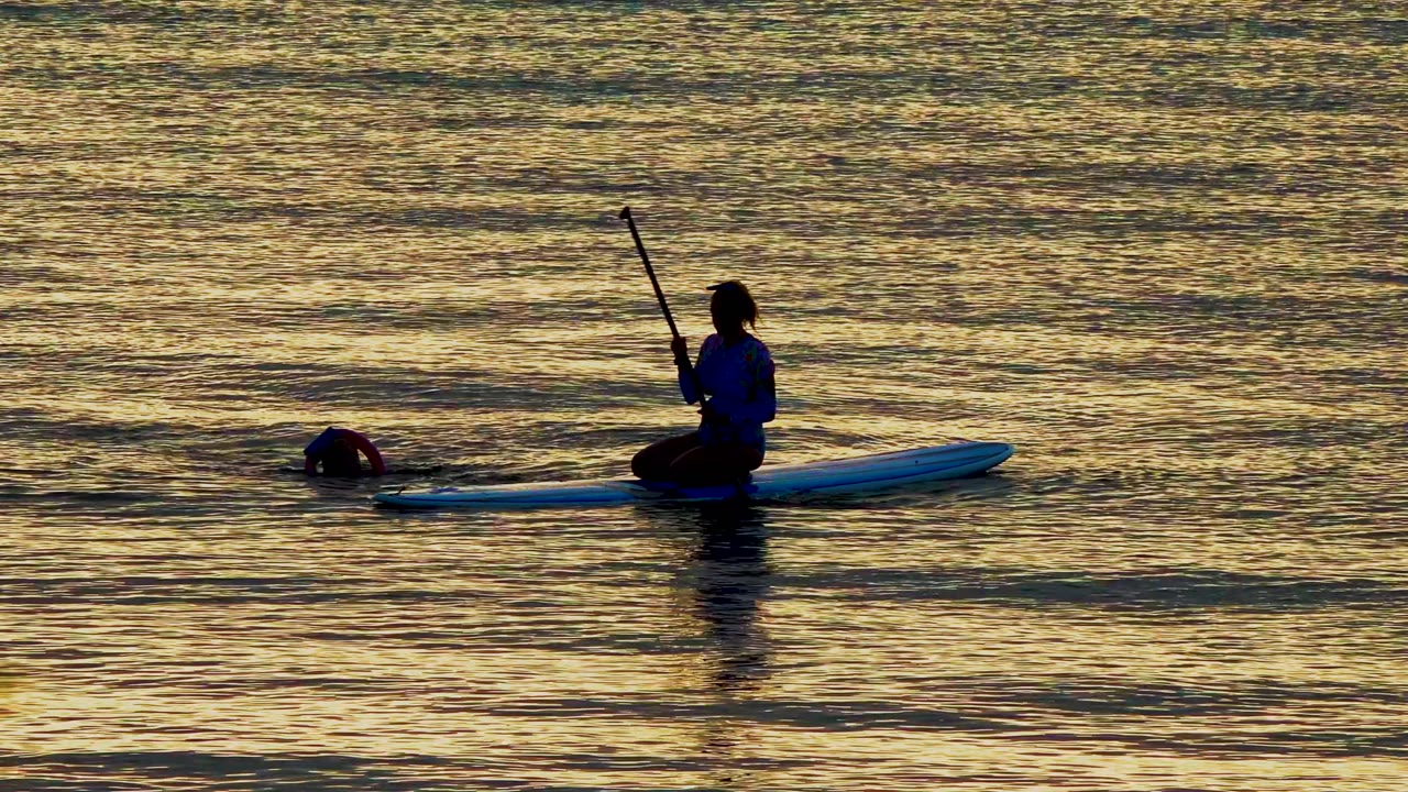 At 0:02 in El Sargento La Ventana Mexico, the paddleboarder sits near the right side of the frame while the swimmer is farther left, partially submerged in the golden water.