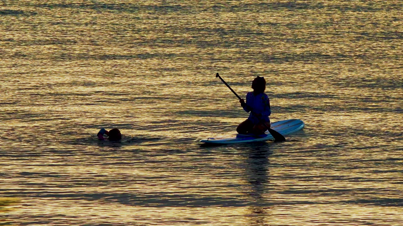 Kneeling paddleboarder near a swimmer in rippled sunset water.