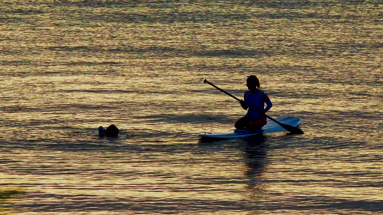 At 0:02 in El Sargento La Ventana, Mexico, the paddleboarder appears on the right side of the frame while the swimmer remains left of the board in dark golden water.