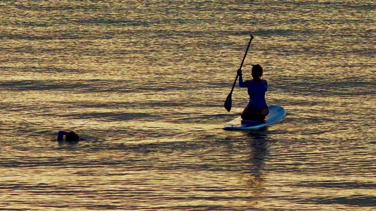 Paddleboarder raises a paddle near a swimmer in golden water