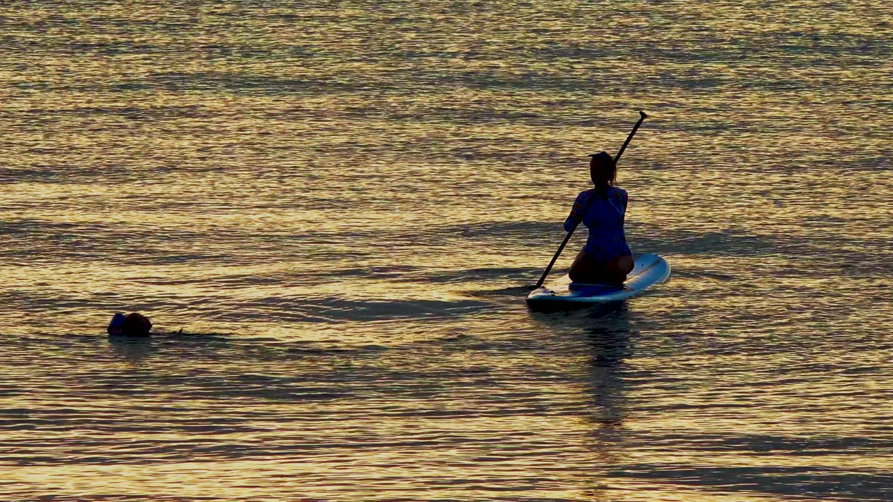 At 0:02 in El Sargento La Ventana, Mexico, the paddleboarder has shifted to the right side of the frame while the swimmer’s head sits left of center in textured golden water.