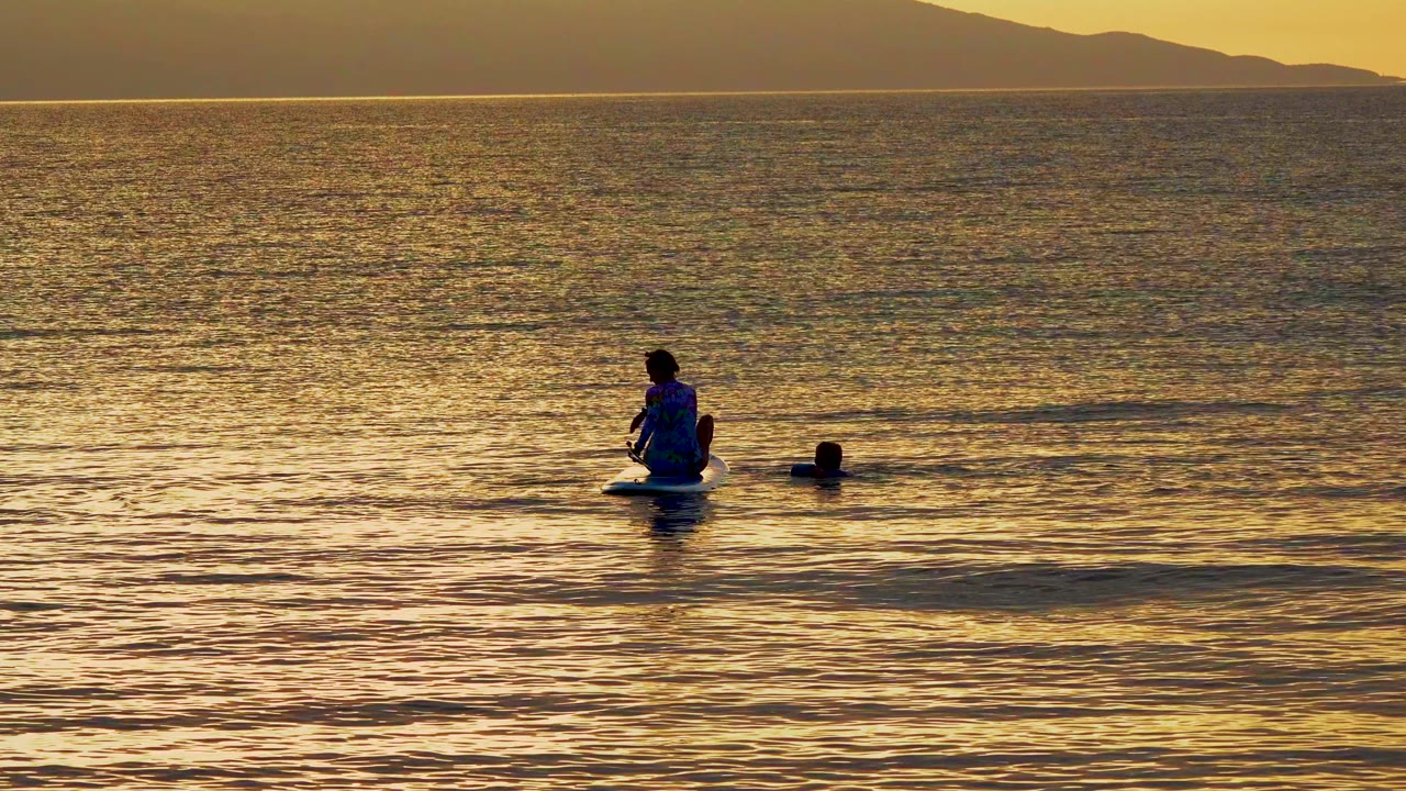 Paddleboarder pauses on calm sunset water with a swimmer nearby.