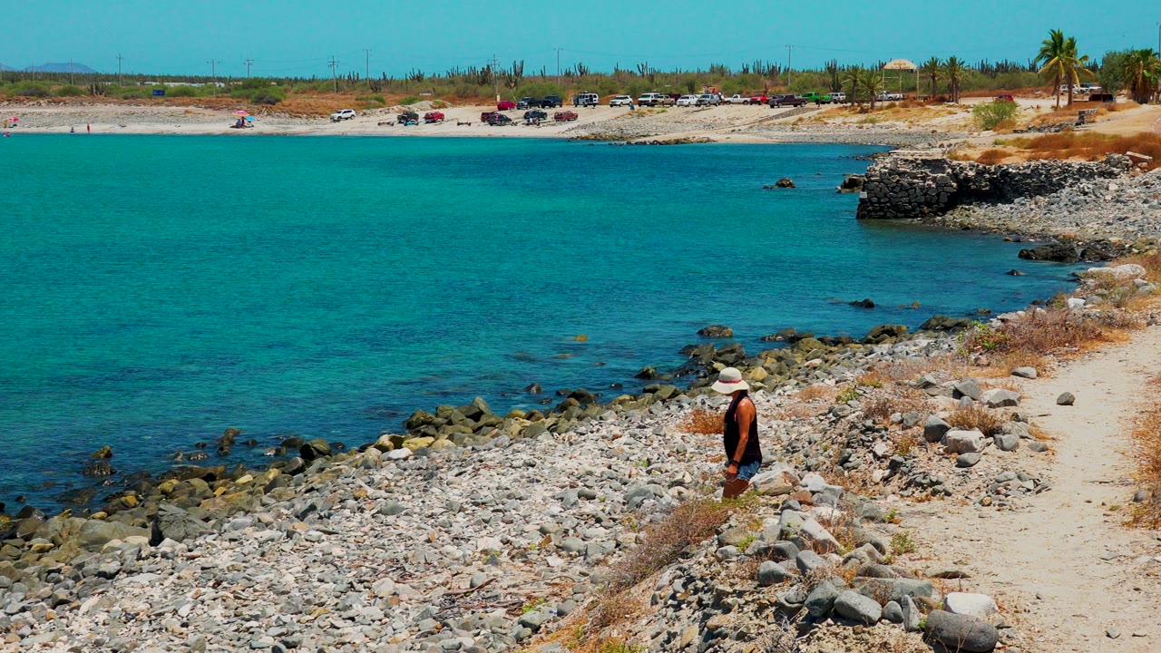 A person in a hat stands on the rocky shore at El Sargento La Ventana Mexico, with clear blue water, a sandy path, and parked cars across the background.