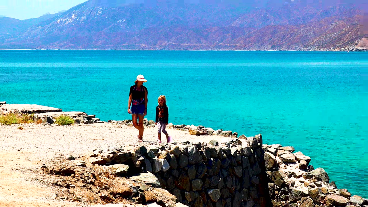 Adult in a white hat and child walk along a pale rocky path in El Sargento La Ventana, Mexico, with turquoise water and brown mountains behind them.