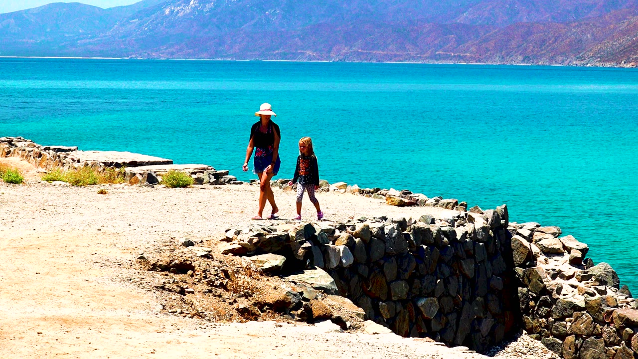 Adult and child continue along a stone-edged coastal path beside blue water.