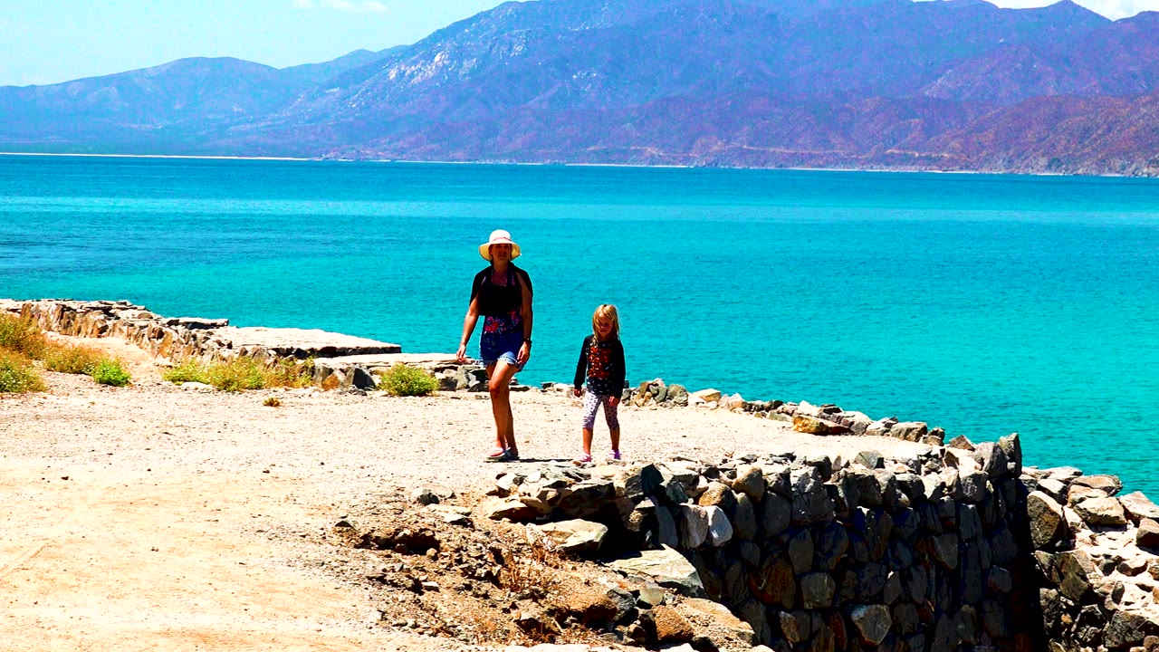 Adult and child walk near a low rock border in El Sargento La Ventana, Mexico, with open turquoise water behind them and mountains across the horizon.