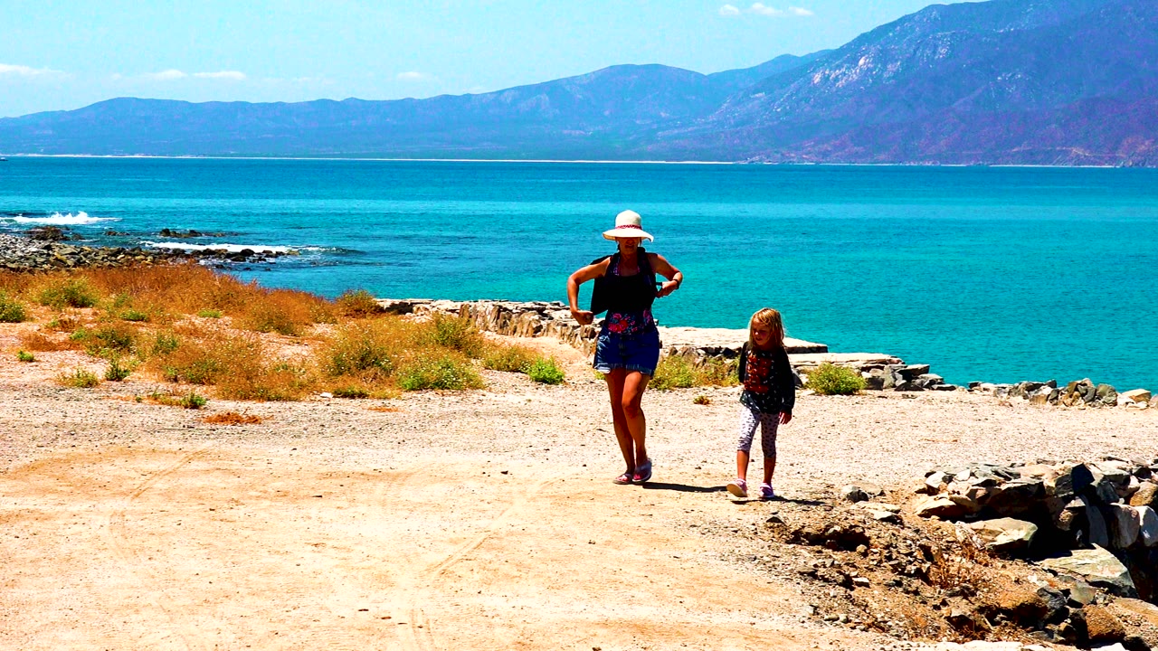 In El Sargento La Ventana Mexico, a woman in a white hat and a child walk across sandy ground with turquoise ocean and mountains behind them.