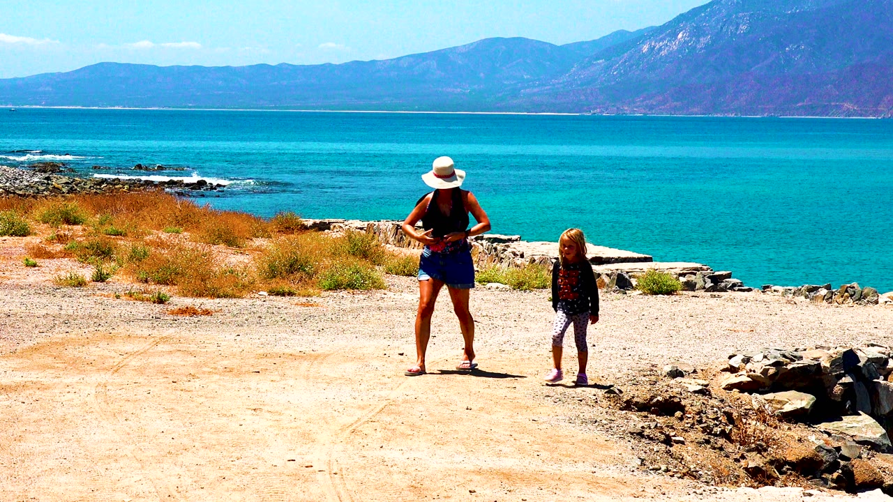 Woman and child paused on sandy ground near rocks and blue water.