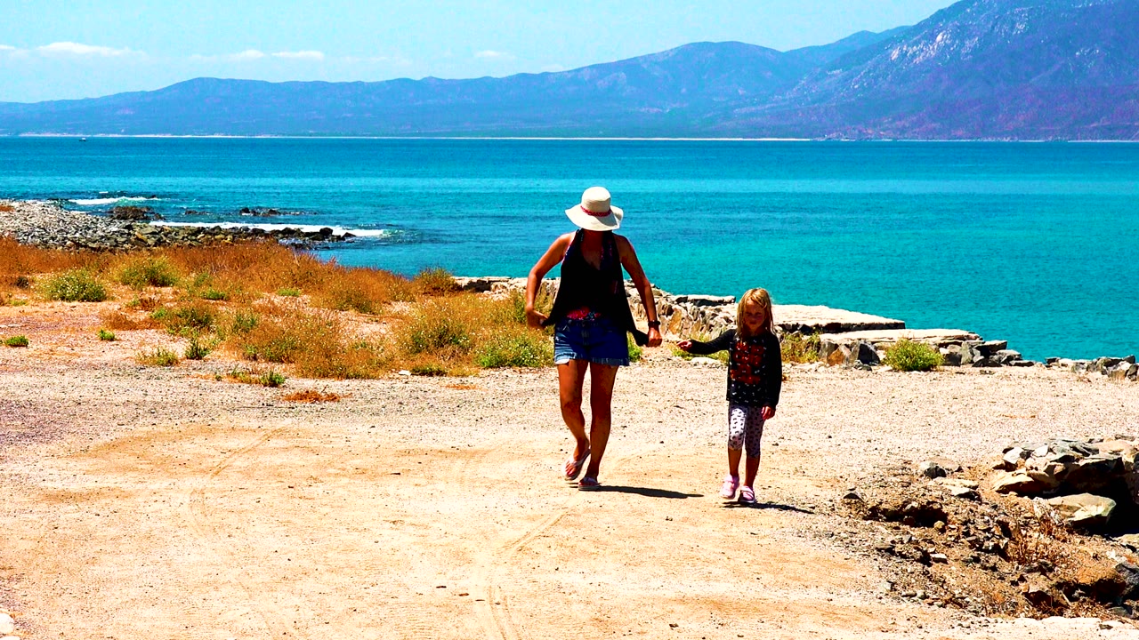 A woman in a sunhat and a child move along the sandy coastal path in El Sargento La Ventana Mexico, with dark rocks at the shore and turquoise sea behind them.
