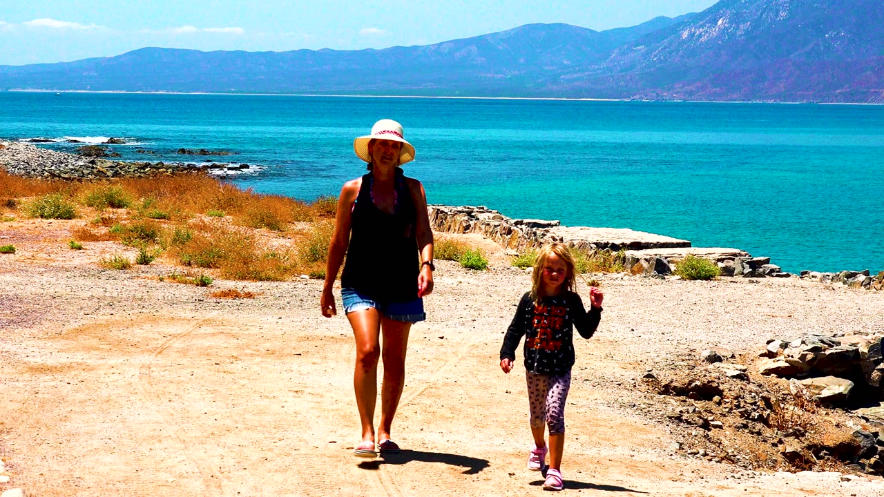 In El Sargento La Ventana Mexico, a woman in a wide sun hat and a young child walk toward camera on a pale dirt path with turquoise water and mountains behind them.