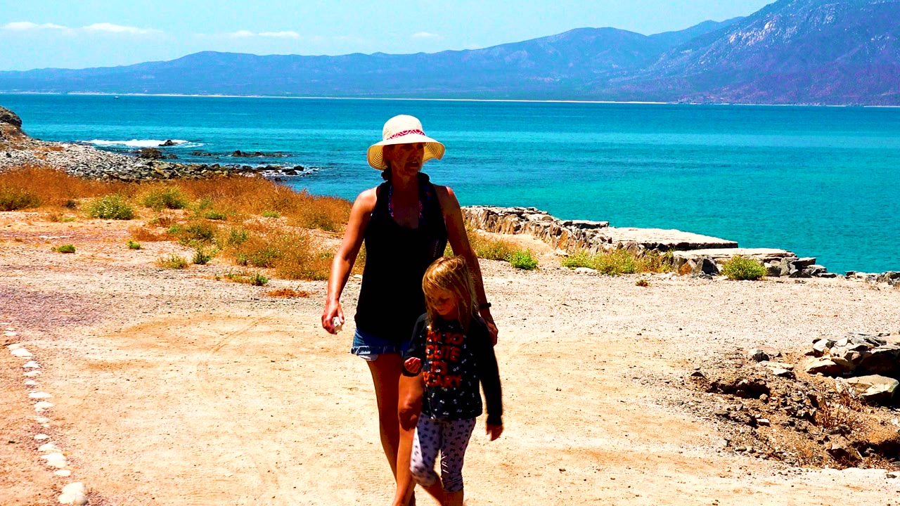 The woman and child move toward the right side of the frame in El Sargento La Ventana Mexico, with the dry path, low rock border, blue ocean, and mountains still visible.