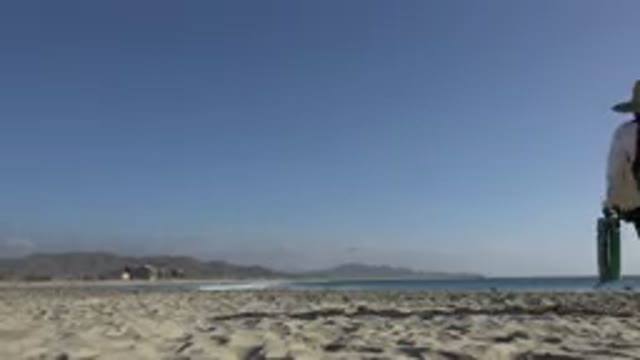 LOS CABOS MEXICO-2017: Person Walking on Beach with Backpack and Hat Near Mountains and Sea