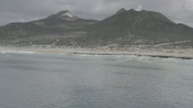 LOS CABOS MEXICO-2020: Waves Crashing On Beach With Mountains And Cloudy Sky